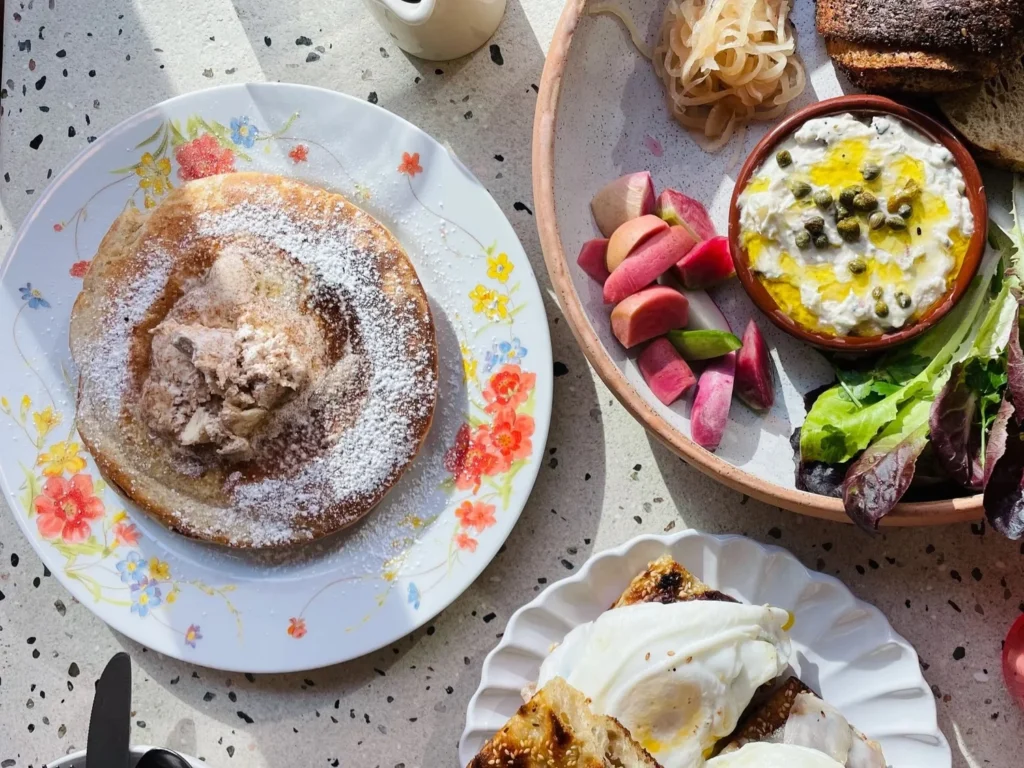 A plate with a large pancake topped with cream, surrounded by sliced apples, salad, and toast.