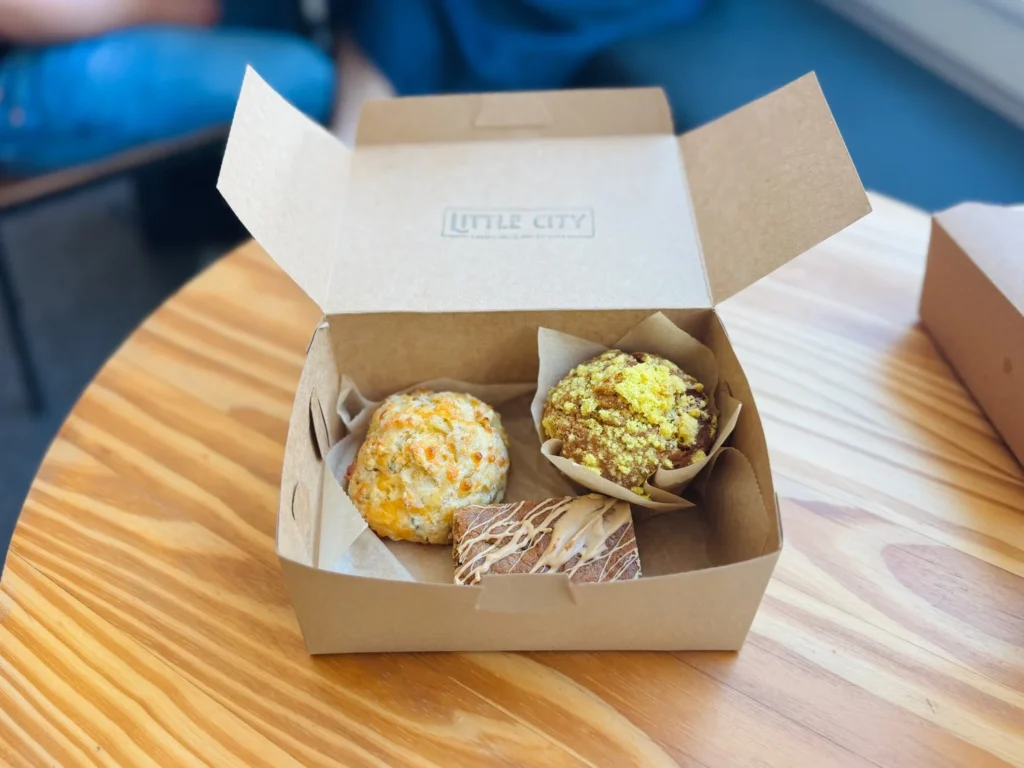 A takeout box contains two baked goods: a cheesy scone and a muffin with yellow topping, on a wooden table.