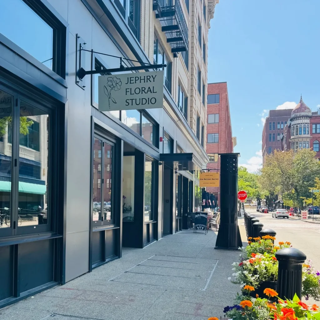 Exterior of Jephry Floral Studio with a sign, sidewalk, and colorful flower planters. Nearby buildings and a stop sign visible.