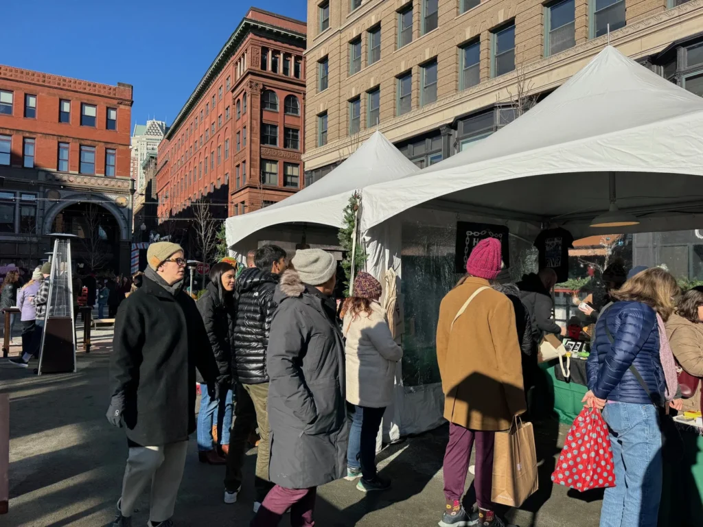 Crowd gathers at a holiday market with tents, shopping bags, and festive decorations in a downtown area.