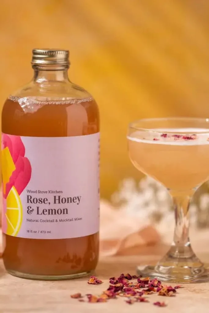 A bottle of Rose, Honey & Lemon mixer next to a cocktail garnished with dried rose petals.