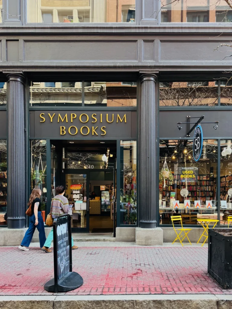Two people walk past Symposium Books, a storefront with a "Open" sign and yellow chairs outside.