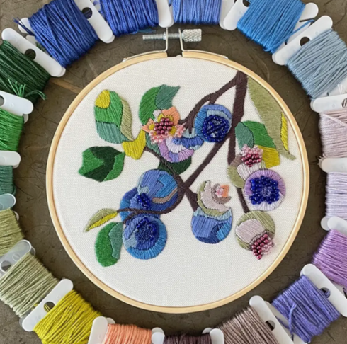 Embroidery hoop displays a floral design with blue berries and green leaves, surrounded by colorful thread spools.