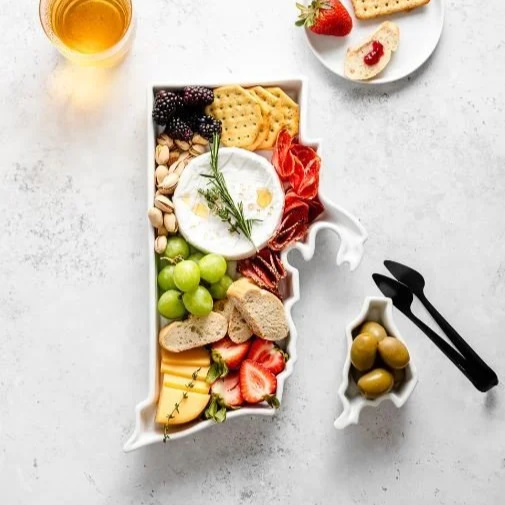 A charcuterie board shaped like a state, featuring cheese, fruits, nuts, and crackers, with a drink nearby.