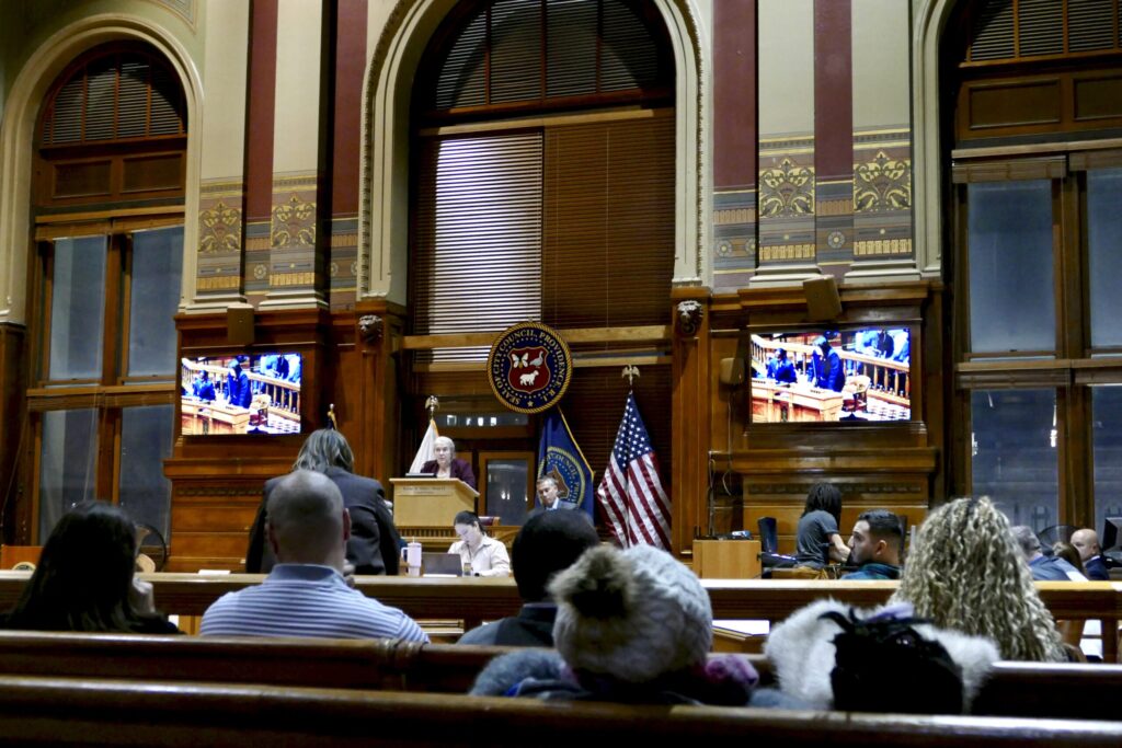 A speaker addresses the assembly in a formal chamber, with audience members seated and monitors displaying proceedings.
