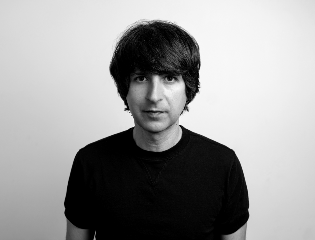 A person with dark, wavy hair stares directly at the camera, wearing a black shirt against a light background.