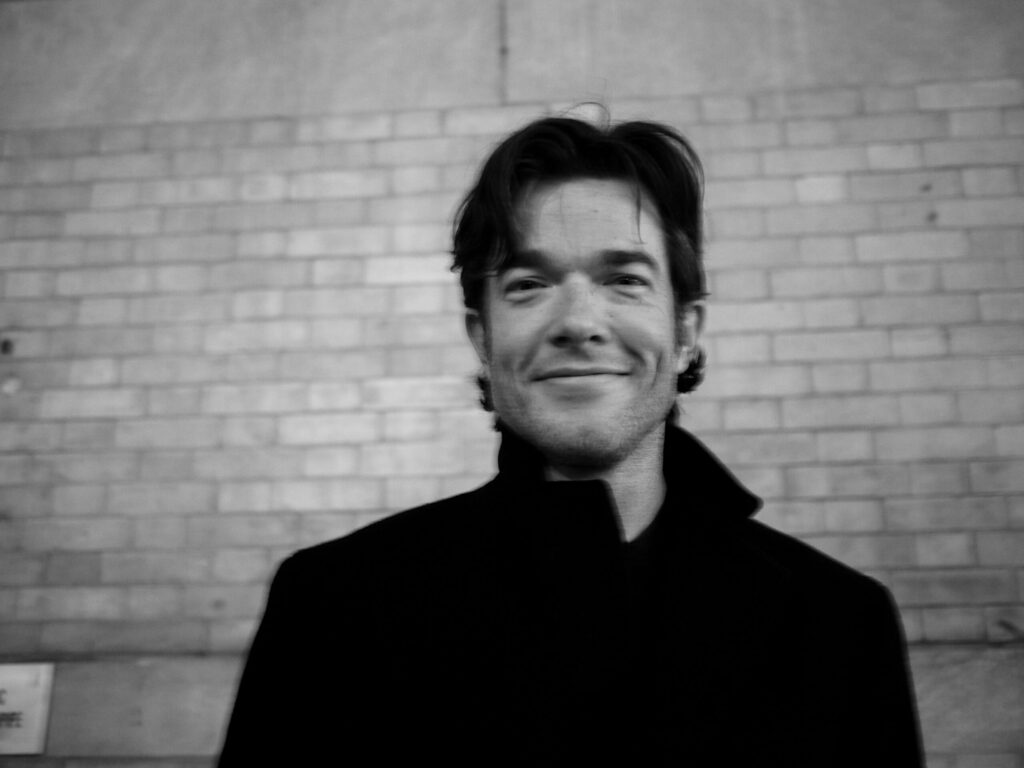 A man with tousled hair smiles, wearing a dark coat against a brick wall background.
