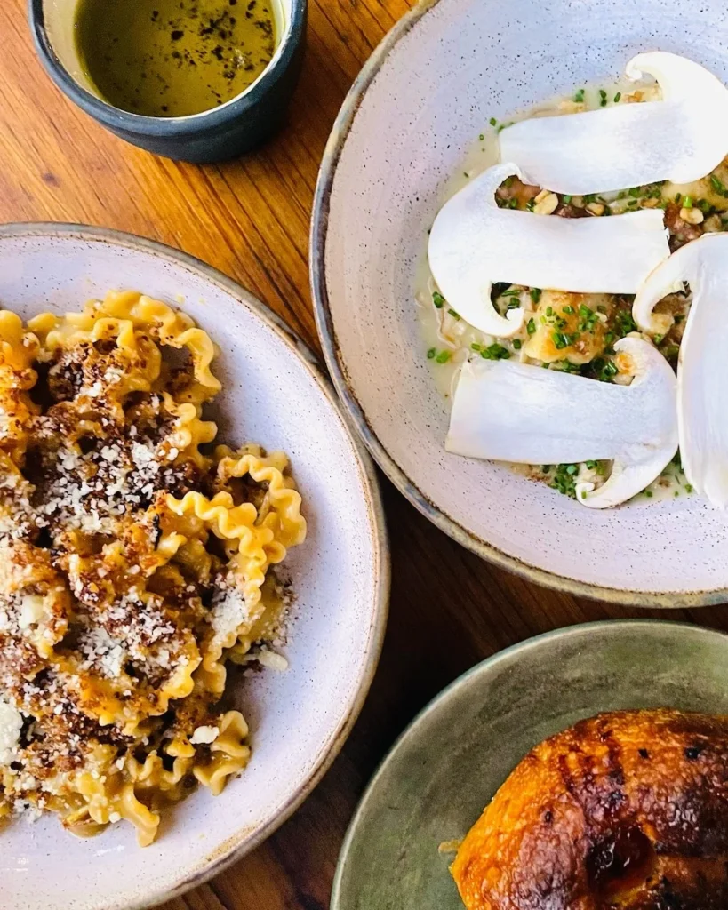 Three plates of pasta dishes with garnishes, a roll, and a small bowl of olive oil on a wooden table.