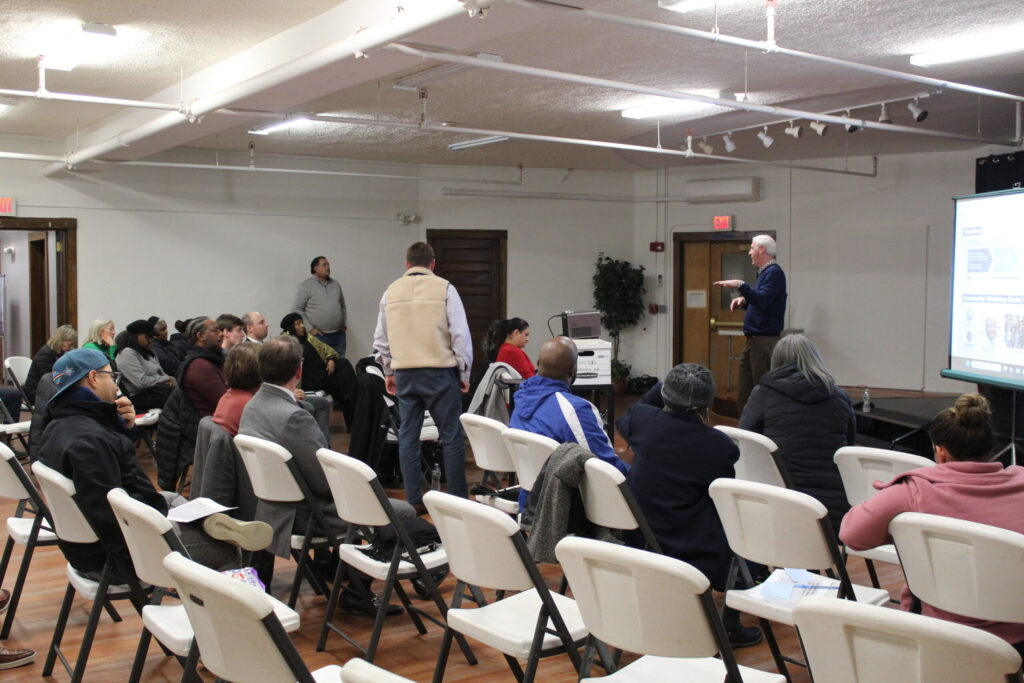 A speaker presents to an audience seated in chairs, with a projector displaying information.