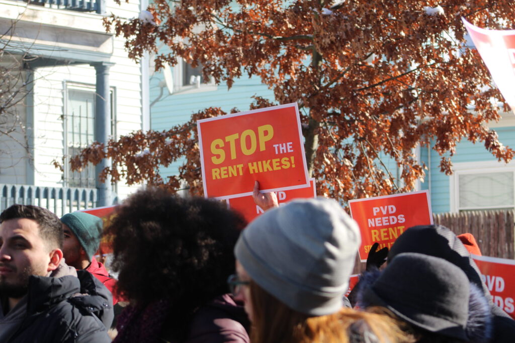 Protesters hold signs demanding to stop rent hikes, with trees and buildings in the background.