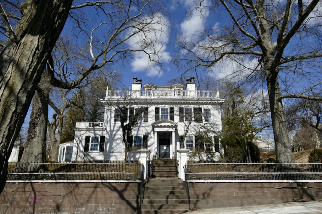 A large white house with a staircase, surrounded by bare trees and a blue sky.