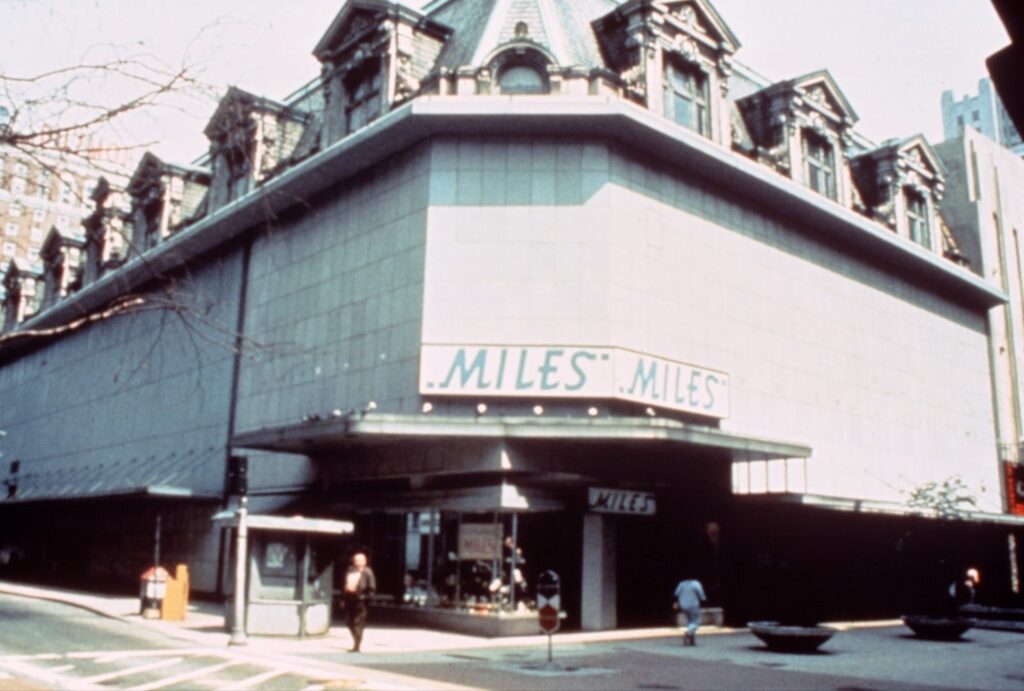 A large building with "MILES" signage, featuring pedestrians and a bus stop nearby.