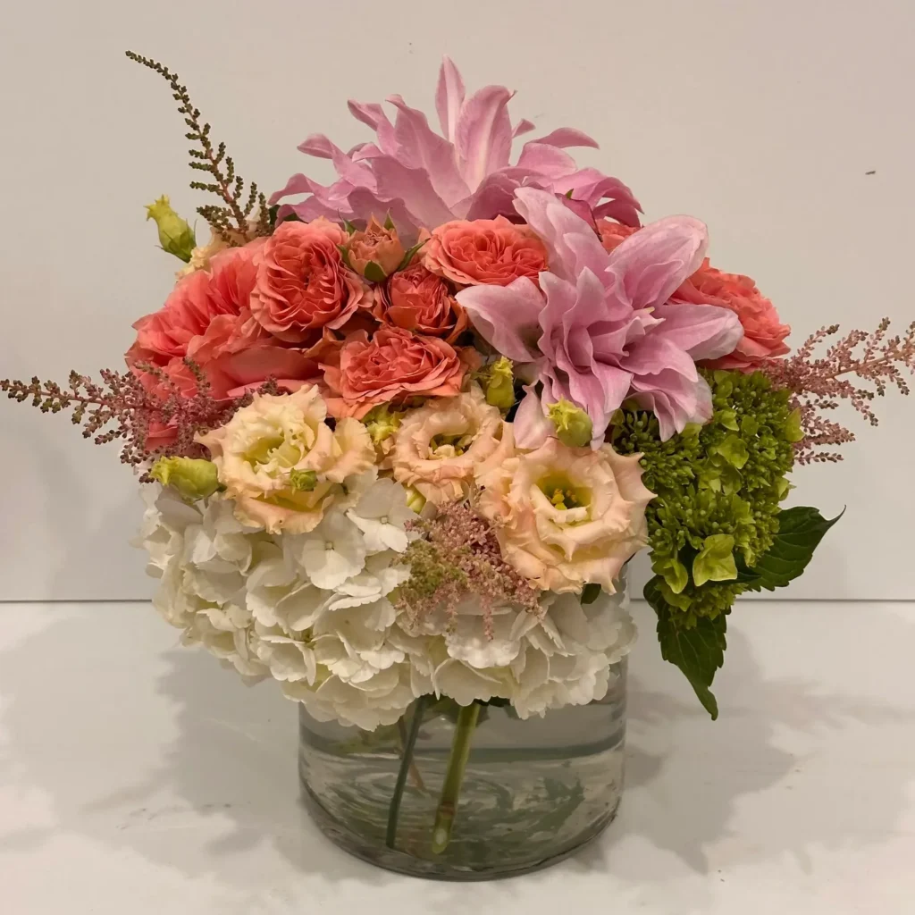 A vibrant floral arrangement featuring pink lilies, coral roses, and white hydrangeas in a clear vase.
