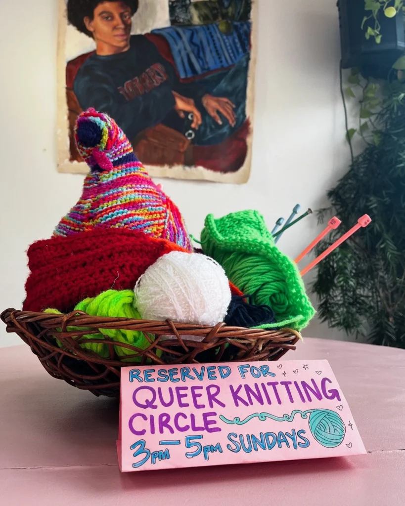 A basket of colorful yarn with a sign for a Queer Knitting Circle, 3-5 PM Sundays. Background features a portrait.