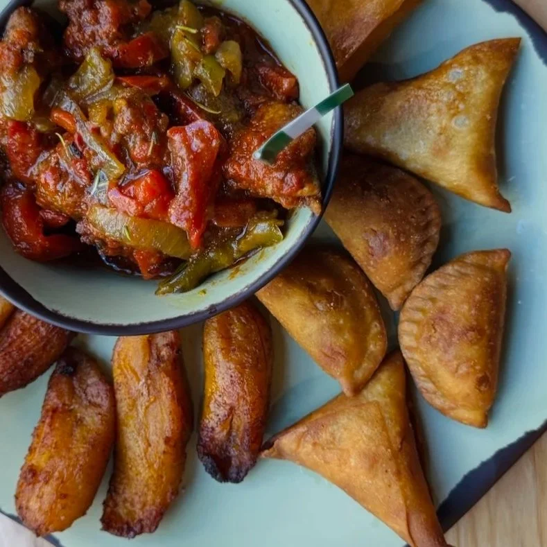 A bowl of spicy stew surrounded by golden fried plantains and triangular pastries.