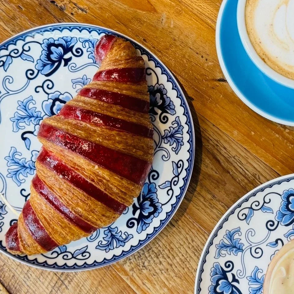 A striped croissant on a decorative plate next to a cup of coffee on a wooden table.