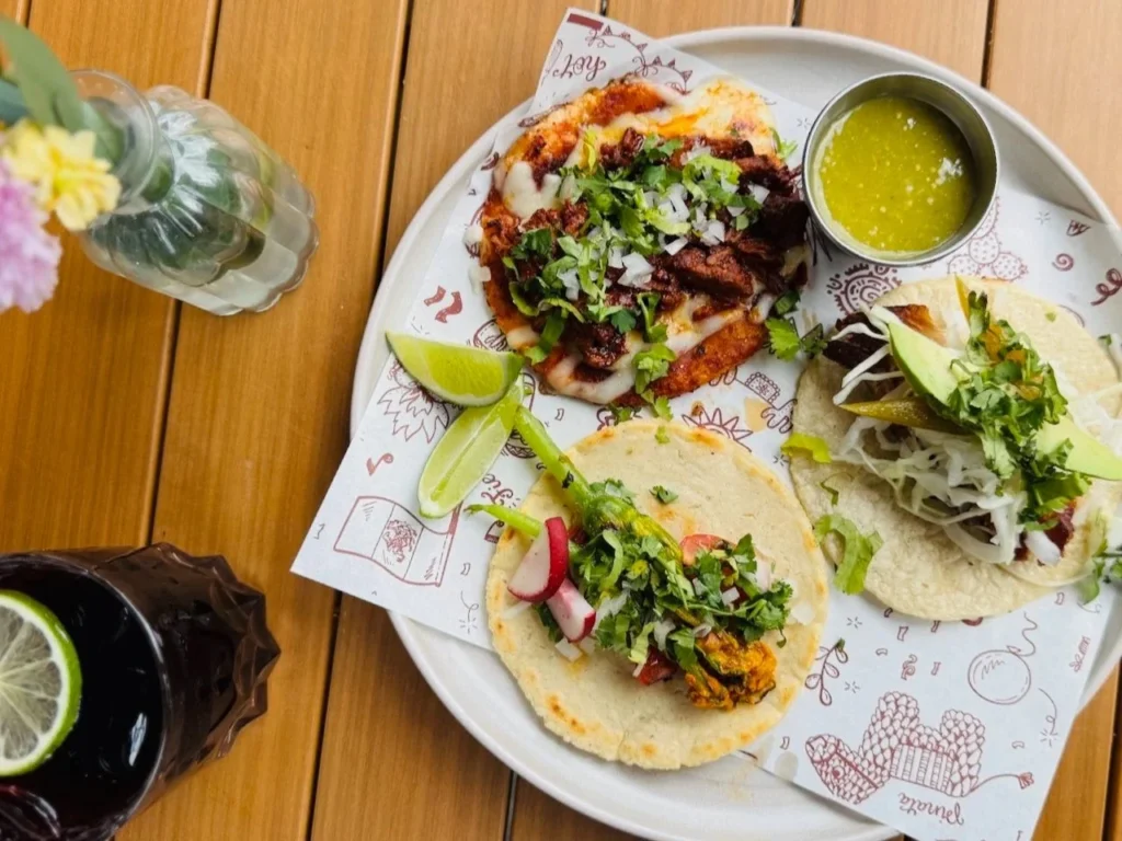 Three tacos with various toppings, lime wedges, and a small bowl of green sauce on a decorative plate.