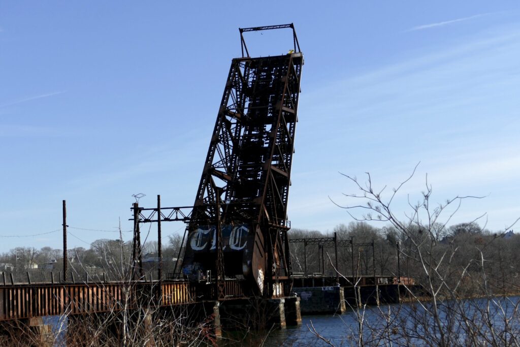 A rusted drawbridge stands raised over a river, surrounded by bare trees and a clear blue sky.