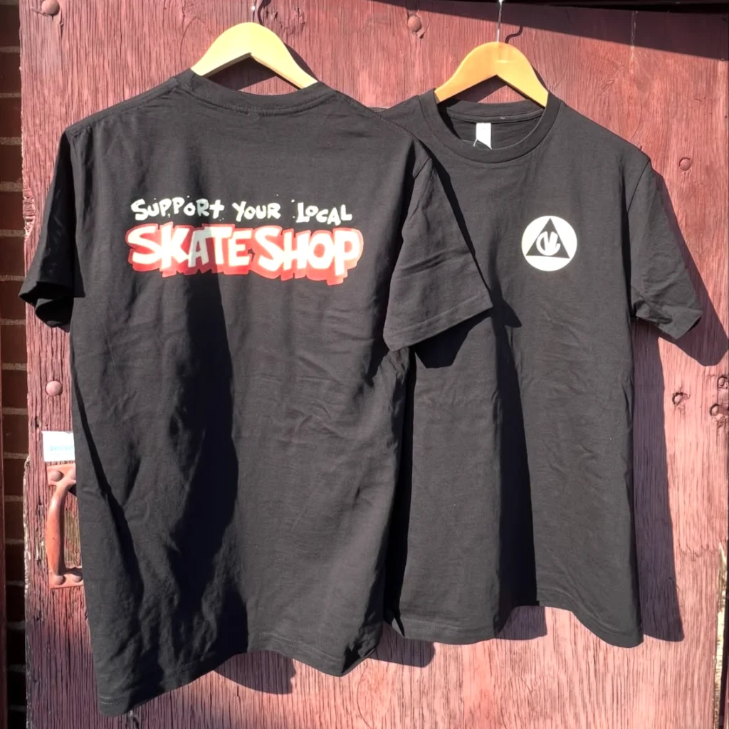 Two black t-shirts hang on a wooden background. One features "Support Your Local Skate Shop" in colorful text, the other has a logo.