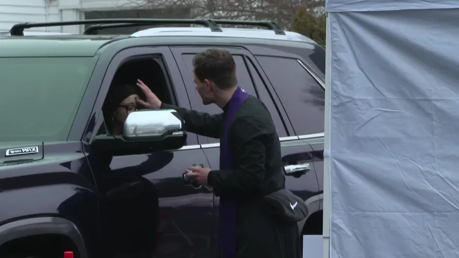 A person in a black coat greets someone inside a vehicle at a drive-thru event.