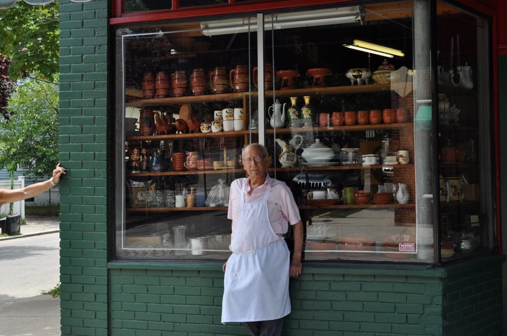 An elderly man in an apron stands by a shop window displaying various pottery and glassware.