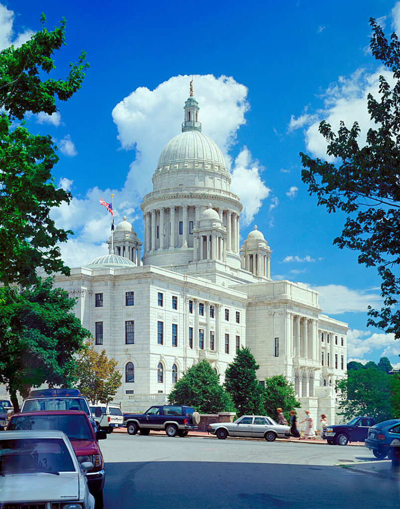 The Rhode Island State House features a large dome under a blue sky with clouds, surrounded by trees and parked cars.