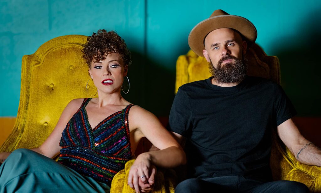 A woman in a sequined top and a man in a black shirt sit together on yellow chairs, both looking serious.