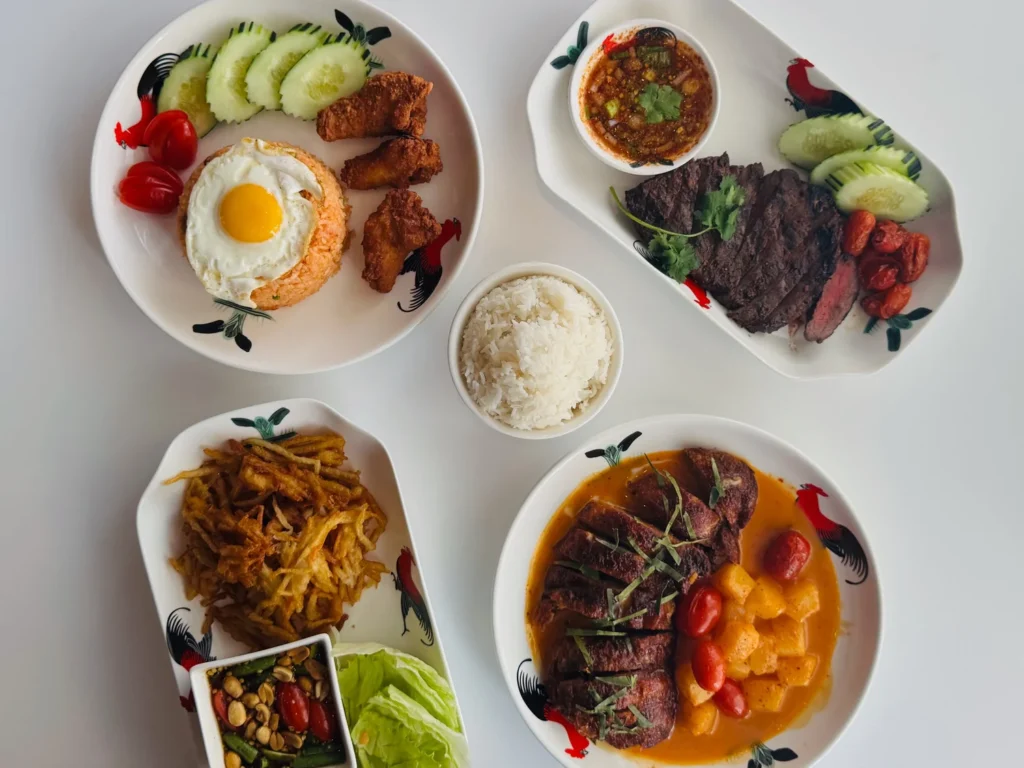 Four plates of food featuring rice, fried chicken, steak, vegetables, and sauces on a white table.