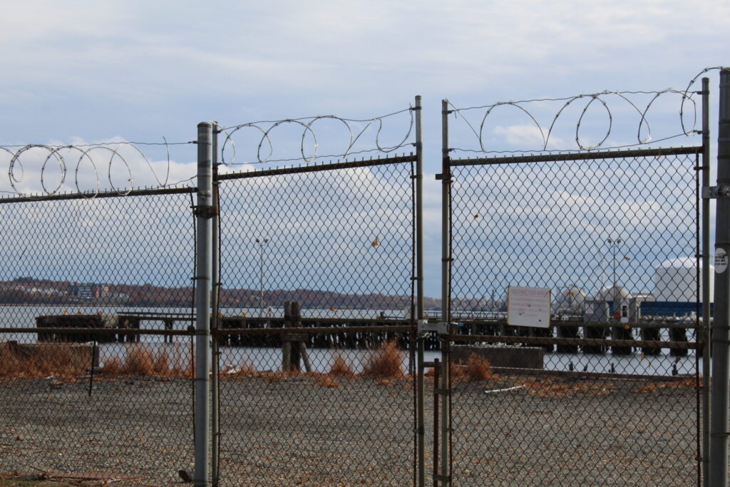 Chain-link fence topped with barbed wire, overlooking a dock and water. A "Restricted Area" sign is visible.