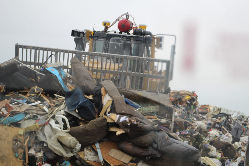 A bulldozer looms over a large pile of mixed waste, including furniture and debris.