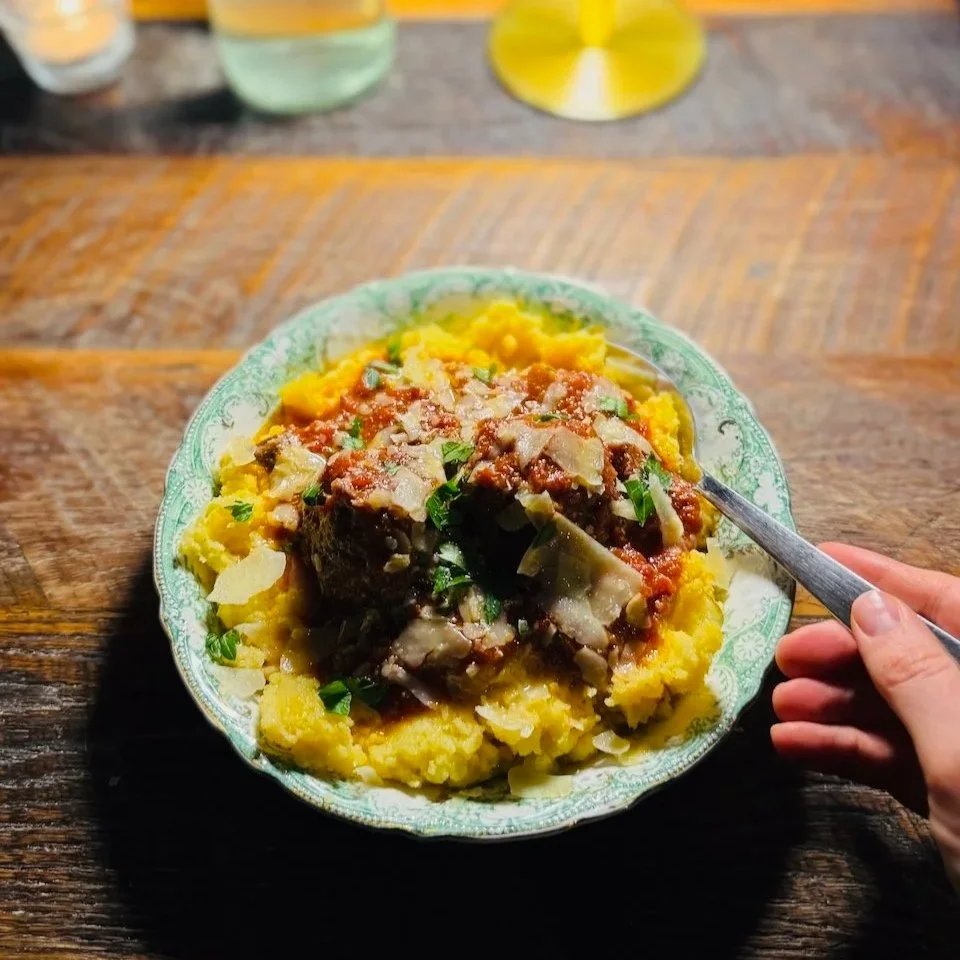 A hand holds a fork over a bowl of polenta topped with meatballs, marinara sauce, and cheese.