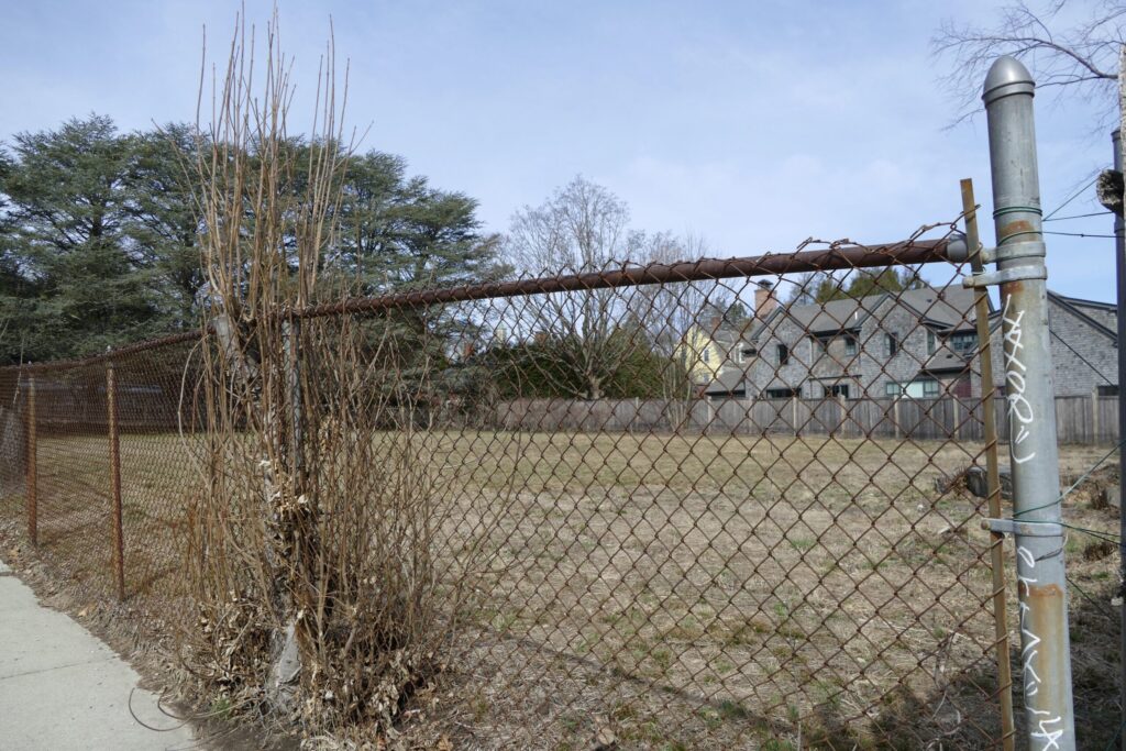 A rusty chain-link fence encloses a barren field, with dry plants and houses visible in the background.