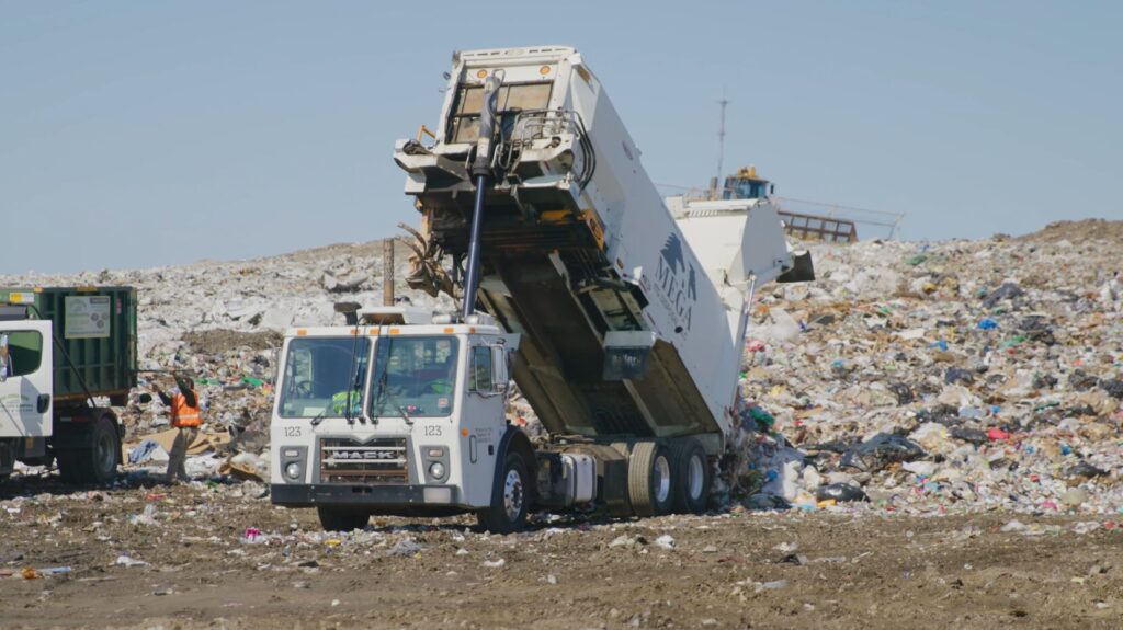 A garbage truck unloads waste at a landfill, with a worker in an orange vest nearby.