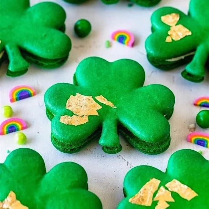 Green shamrock-shaped macarons with gold accents, surrounded by colorful candies and rainbow decorations.