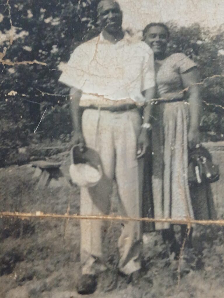 A man and woman stand together outdoors, both smiling. The man holds a hat, and the woman carries a handbag.