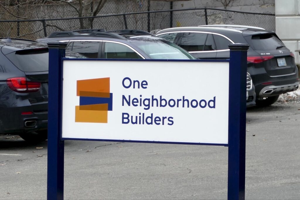 Sign for "One Neighborhood Builders" in front of parked cars.