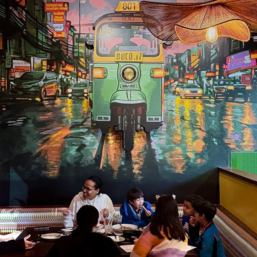 A group of diners enjoys a meal at a restaurant with a colorful mural of a green tuk-tuk.