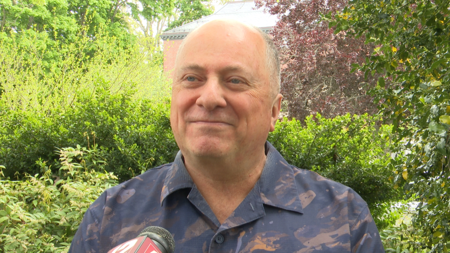 A man smiles while standing outdoors, surrounded by greenery and trees.