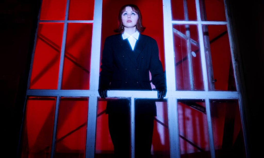 A person in a black suit stands at a window, gazing out against a red backdrop.