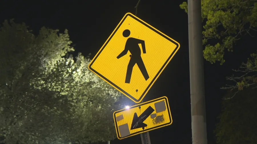Yellow pedestrian crossing sign with a walking figure, illuminated at night, above a directional arrow sign.