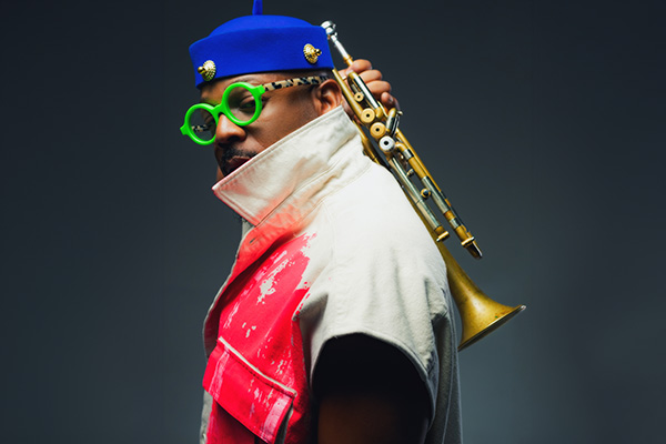 A man with a trumpet rests on his shoulder, wearing a blue hat and green glasses, in a stylish outfit.