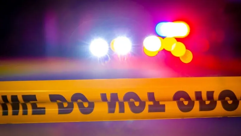 Yellow police tape reads "DO NOT CROSS" in front of flashing emergency lights.