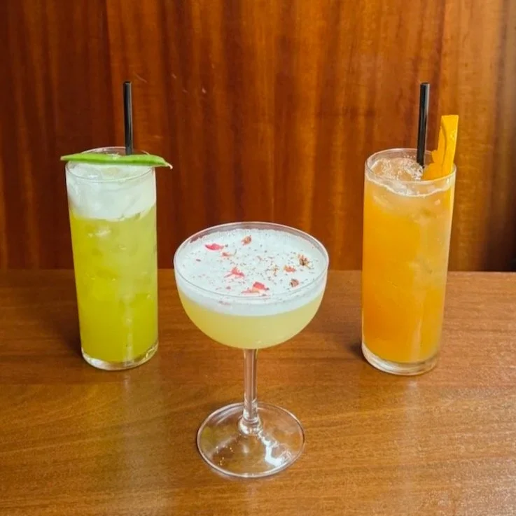 Three colorful cocktails on a wooden table: a green drink, a yellow drink with rose petals, and an orange drink.