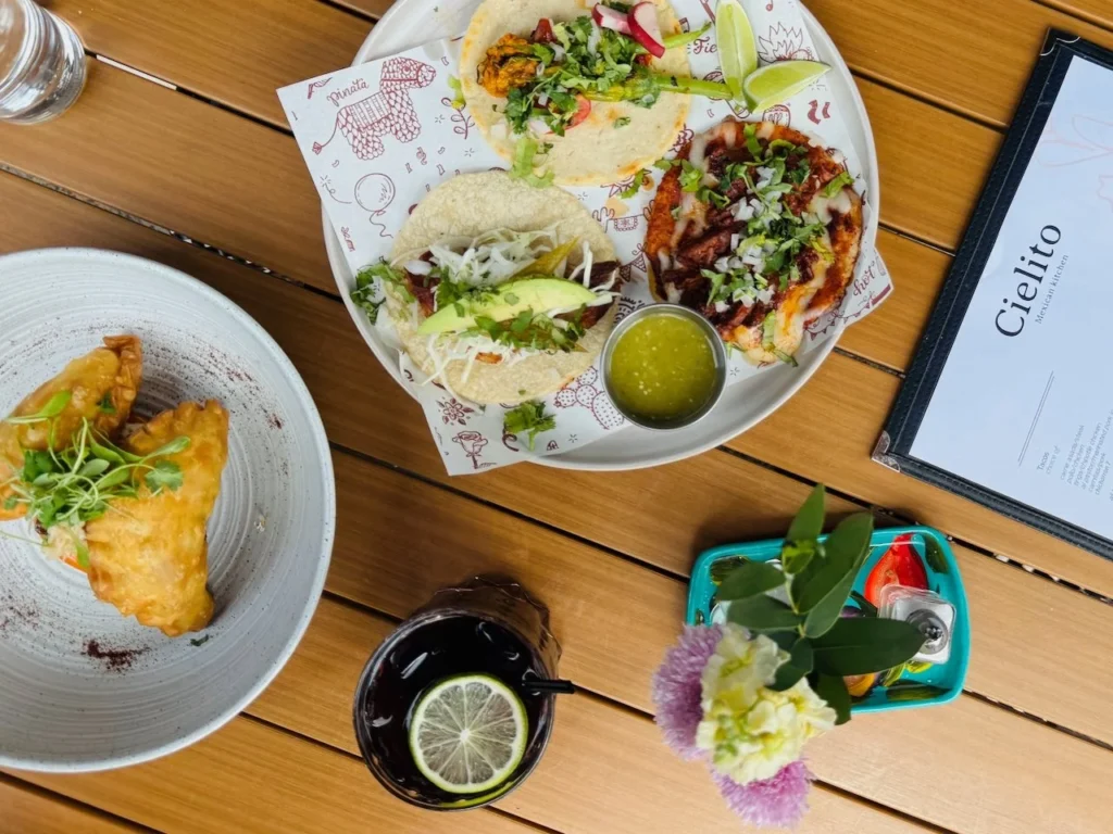 Plates of tacos and empanadas on a wooden table, with a drink and a small vase of flowers.