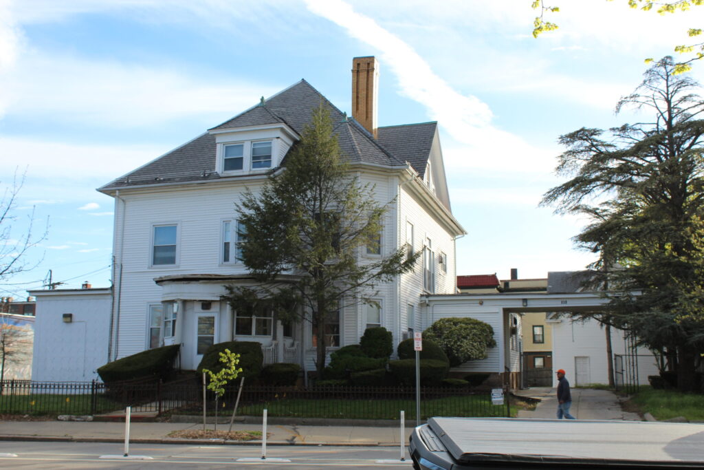 Large white house with a steep roof, surrounded by trees and shrubs; a person walks nearby.