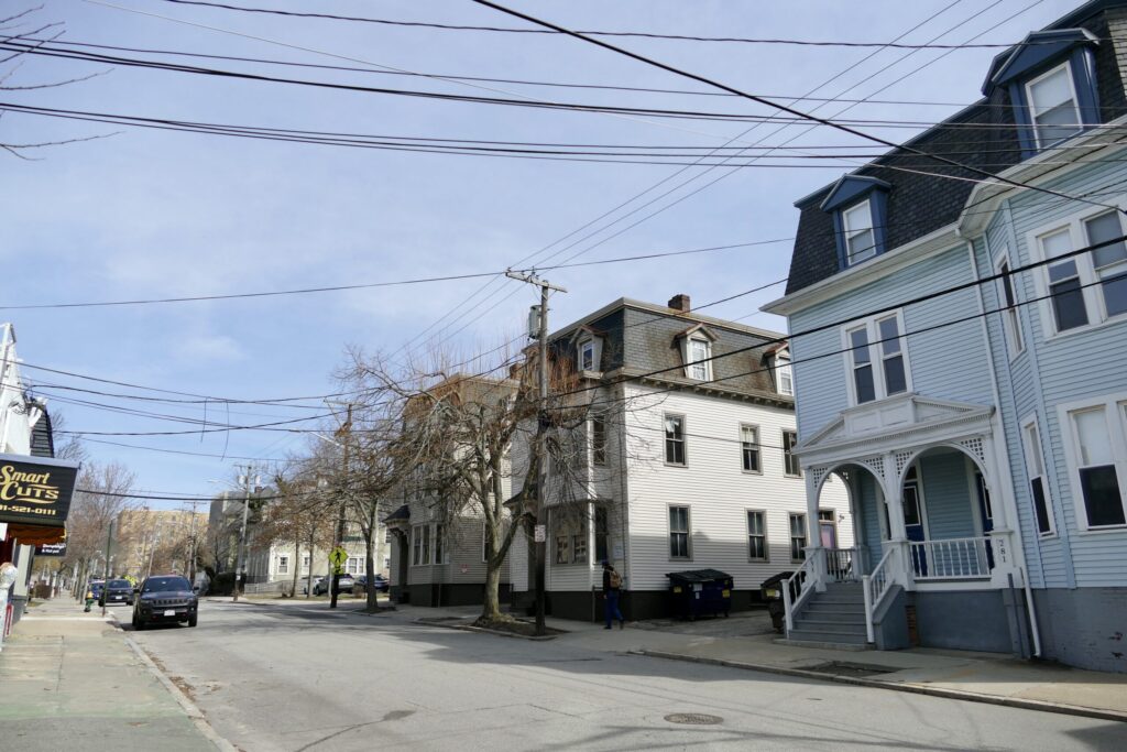 Street view featuring residential buildings, utility poles, and parked cars under a clear sky.