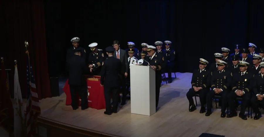 A ceremony with officials at a podium, presenting awards to uniformed personnel seated on stage.