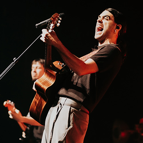 A musician passionately sings and plays guitar on stage, with a band member in the background.