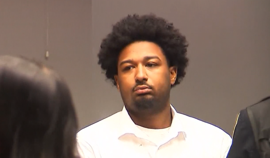 A man with curly hair looks serious while seated in a courtroom setting.