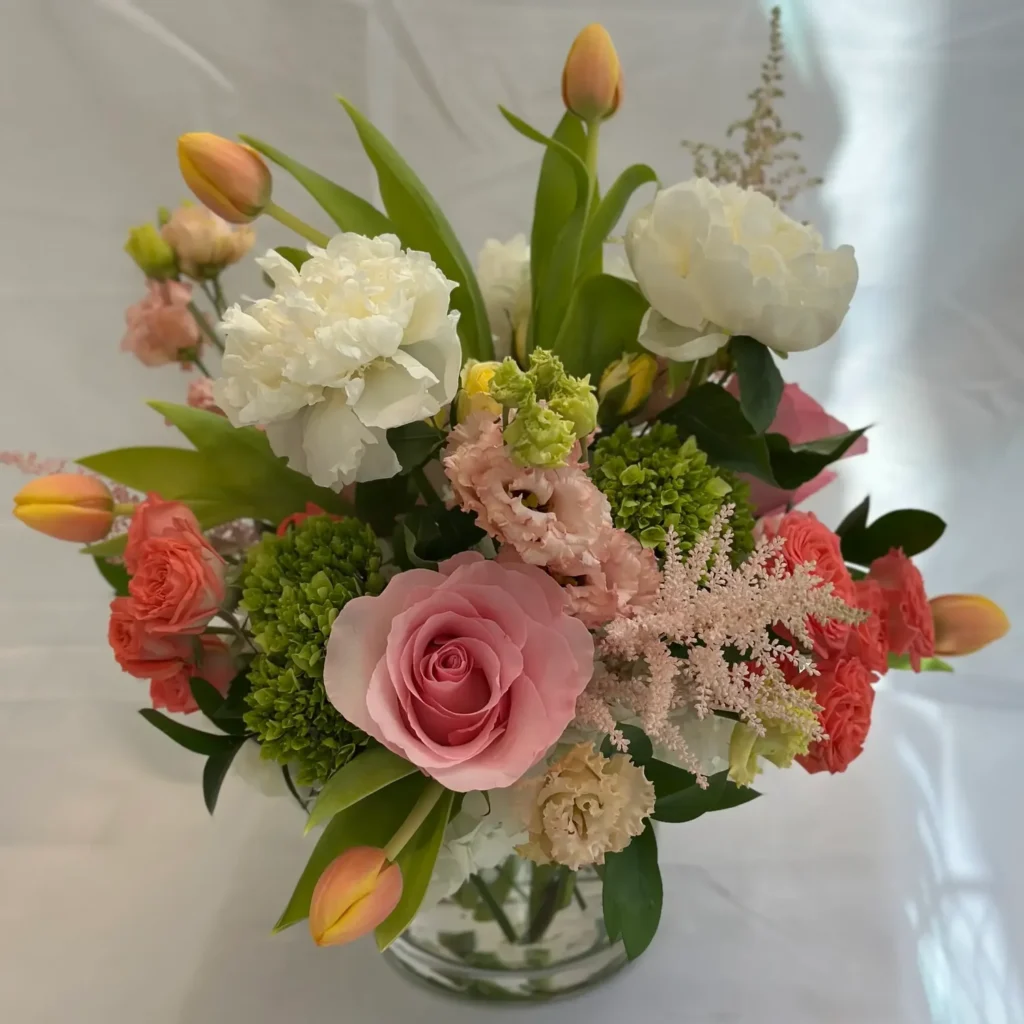 A vibrant bouquet featuring pink roses, white peonies, and green hydrangeas in a glass vase.